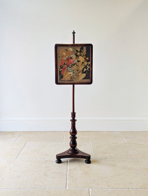 Antique William IV rosewood pole screen featuring a cushion-moulded frame enclosing a parrot needlepoint panel, set on a turned double-baluster stem and triform base.