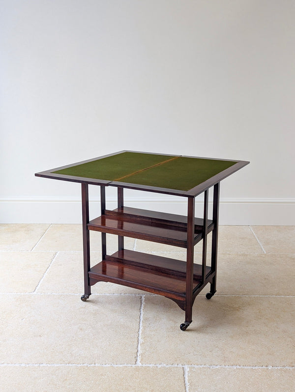 Antique early 20th century mahogany card table featuring a swivelling fold‑over green baize top and a tiered bookshelf base on brass and ceramic castors.