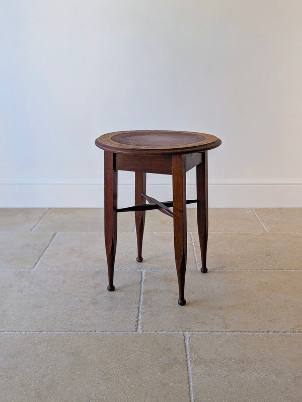 Antique Edwardian Arts & Crafts oak occasional table with a circular oxblood panel top, square-tapered flared legs, swept X-stretcher, and turned ball feet.