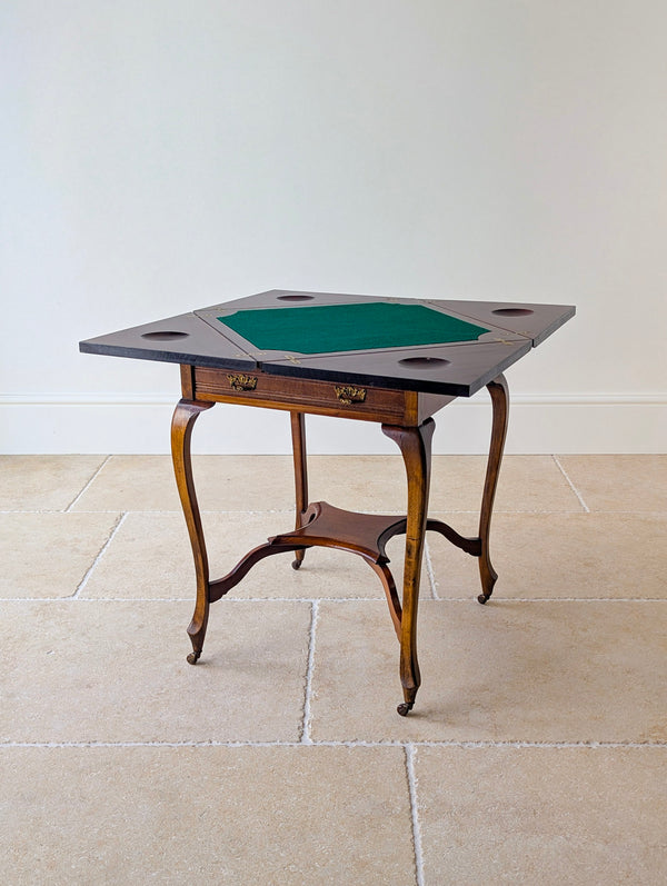 Antique Edwardian mahogany envelope card table featuring a quartered top, single frieze drawer, cabriole legs, shaped X-form stretcher, and brass and ceramic castors.