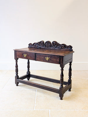 Antique Jacobean revival Victorian oak side table with single plank top, heavily carved rear cresting, two frieze drawers and turned legs united by a stretcher frame.