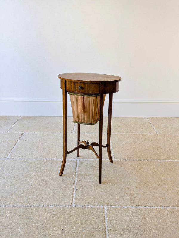 Antique Edwardian mahogany work table featuring a quarter‑veneered oval top and single drawer with lower sliding work basket on slender line‑inlaid legs with serpentine X‑stretcher.
