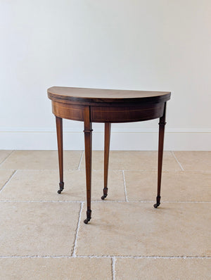 Antique Edwardian mahogany demilune fold-over card table featuring geometric crossover inlay, a green playing surface, and square tapered legs on castors.