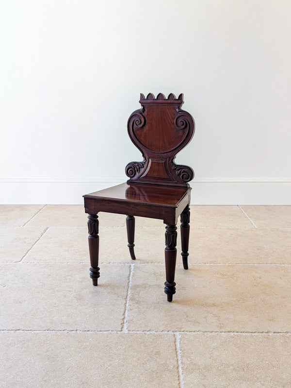 Antique William IV mahogany hall chair featuring a castellated cresting rail, cartouche backboard, solid seat and turned legs with lotus-leaf collars.