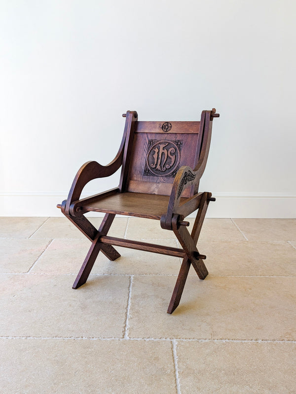 Antique Edwardian Gothic oak Glastonbury chair with a relief-carved IHS Christogram backboard, swept foliate arms, and solid seat, raised on an X-frame support.  