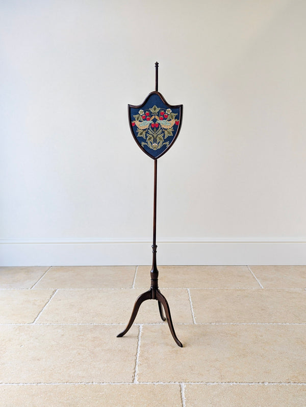 Antique George III mahogany tripod pole screen with an urn finial, shield-shaped frame housing a William Morris needlepoint panel, and down-swept legs.